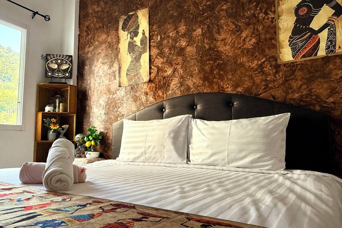 A neatly made bed with white linens, a quilt with a colorful pattern, a wooden nightstand with decorative items, and a large window with a view of trees.