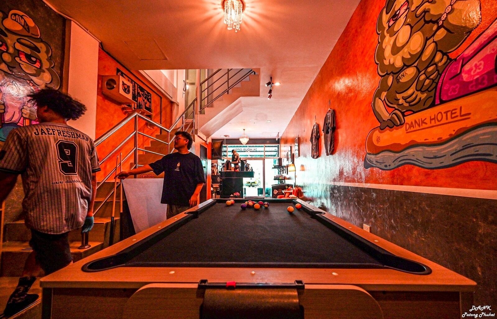 Une table de billard dans une pièce ornée d’une fresque murale, avec un escalier et une enseigne indiquant « HÔTEL DANX ».
