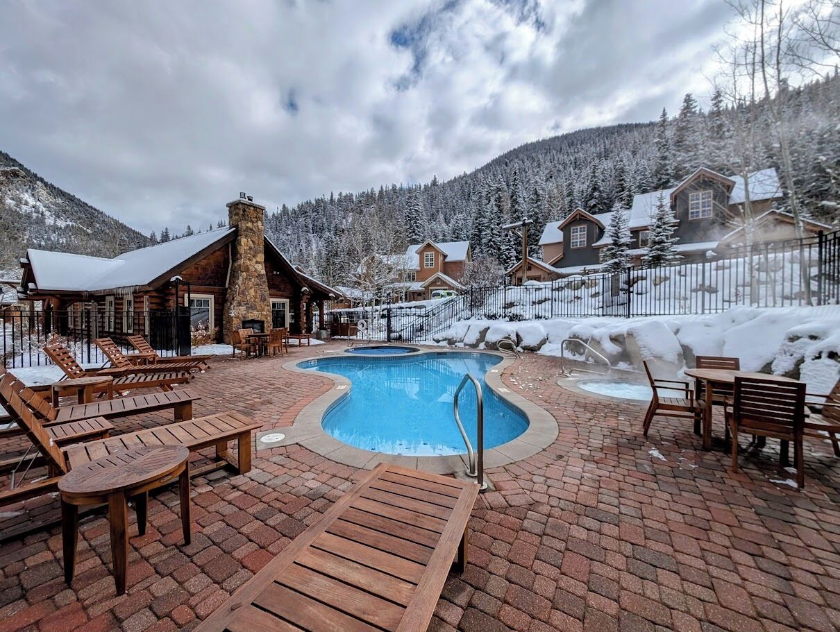 Un espace piscine doté de chaises longues et d’une cheminée en pierre, entouré d’arbres et de montagnes couverts de neige.