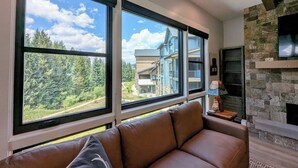 A modern living room with a leather sofa, large windows offering a view of a building and trees, and a stone fireplace.