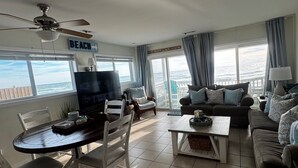 A living room with a TV, dining table, and chairs, a sofa, and a view of the ocean. - Oceanfront Ground Floor End-Unit Condo w/pool -
Shamrock Sands M1 @ The Riggings (Kure Beach)
