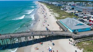 A beach with a pier, beachgoers, and a building with a blue roof. - Oceanfront Ground Floor End-Unit Condo w/pool -
Shamrock Sands M1 @ The Riggings (Kure Beach)