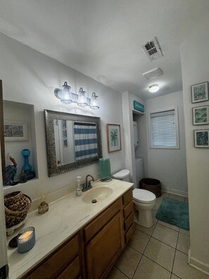 A bathroom with a sink, mirror, and toilet. There are decorative items on the counter and a window with blinds.