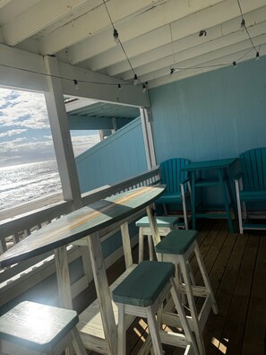A covered outdoor seating area with a view of the ocean, wooden tables and chairs, and a blue wall.