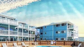 A pool area with a wooden deck, lounge chairs, and a view of a building with multiple balconies. - Oceanfront Ground Floor End-Unit Condo w/pool -
Shamrock Sands M1 @ The Riggings (Kure Beach)