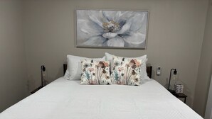 A neatly made bed with a floral pillow set, a large floral artwork above the bed, and bedside tables with lamps and a clock. - Clinton Cabins #2 (Clinton)