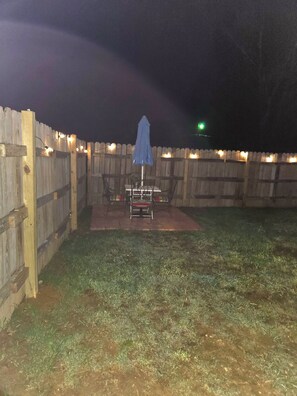 Un espace extérieur clos, aménagé avec une table et des chaises, un parasol bleu et des guirlandes lumineuses.