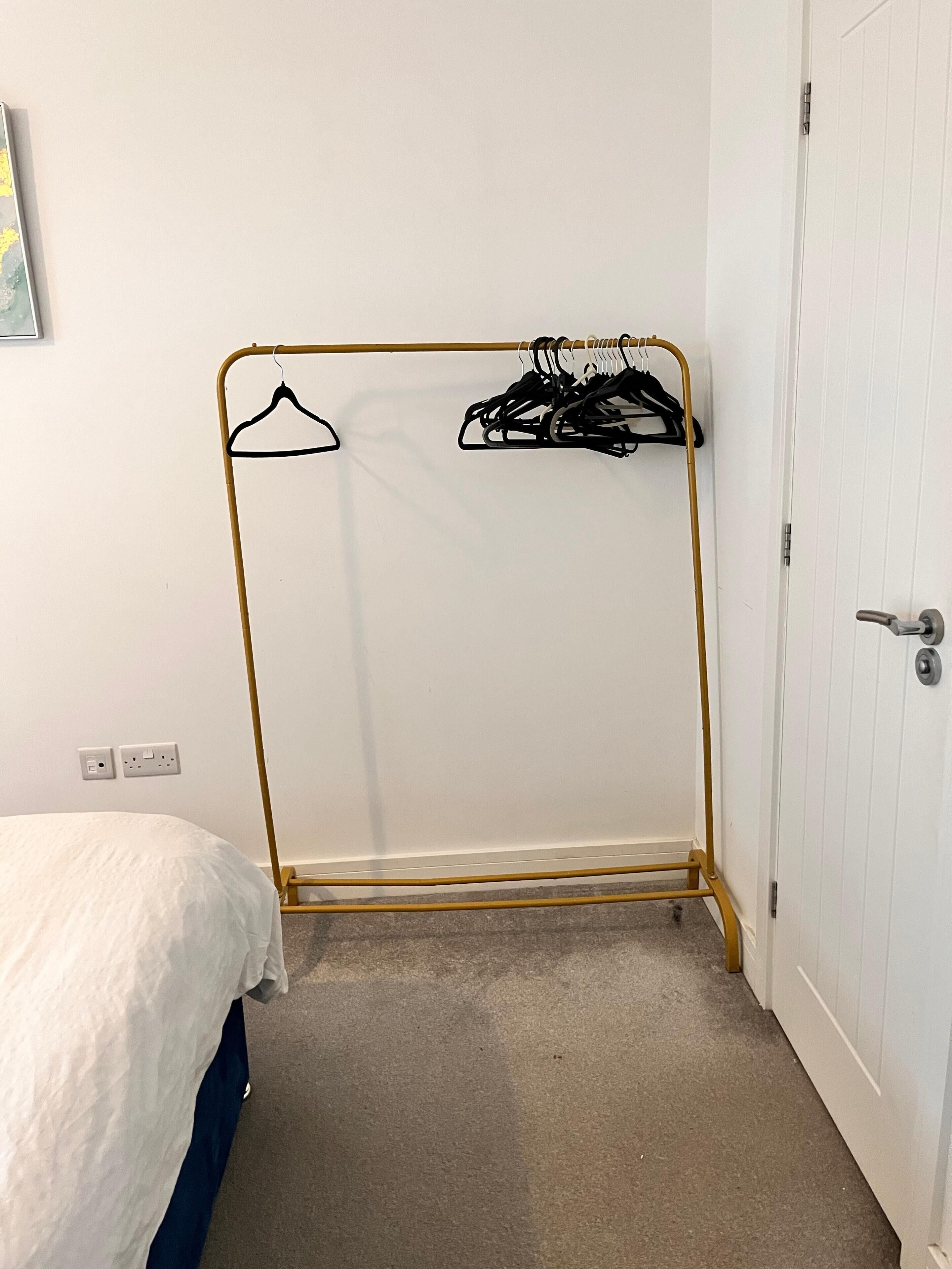 Une pièce avec un lit, un portant métallique sur lequel sont suspendus des vêtements, une porte blanche et un sol recouvert d’une moquette.