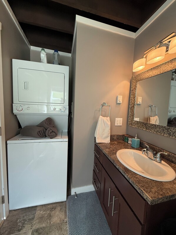 A bathroom with a built-in washer and dryer, a sink with a granite countertop, and a large mirror with integrated lighting.