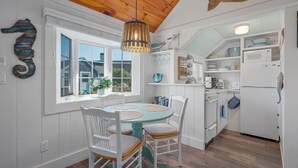 A kitchen with white cabinets, a wooden ceiling, and a dining area with a round table and white chairs. - Catch a Cape Escape at By The Sea (South Yarmouth)