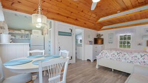 A cozy cabin-style room with a wooden ceiling, a dining area with white chairs and a table, a bed with floral bedding, and a television on a dresser.