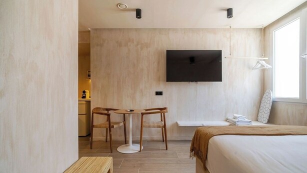 Une chambre d’hôtel moderne équipée d’un lit, d’un téléviseur fixé au mur, d’un petit bureau avec des chaises et d’une fenêtre laissant entrer la lumière naturelle.