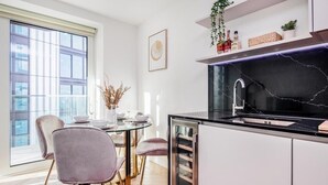 Una cocina moderna con encimera negra, armarios blancos y un refrigerador con puerta de vidrio. Hay un comedor con una mesa redonda y sillas.
