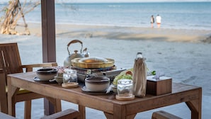A wooden table set with various dishes and a portable stove, overlooking a beach with people in the distance.
