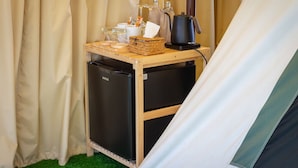 A compact kitchenette with a refrigerator, a coffee maker, and a small table with a basket, glass, and a tissue box.