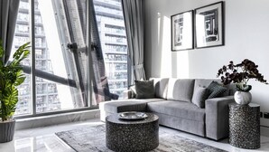 A modern living room with a grey sofa, a round coffee table, and a large window with curtains. - Premium Corner Apt 1BD in JONE w Canal View (Dubai)
