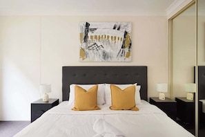 Une chambre d’hôtel avec un grand lit, deux tables de chevet avec des lampes, une grande peinture abstraite au mur et un miroir sur le côté droit.