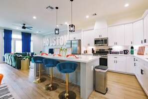 A modern kitchen with white cabinets, a central island with bar stools, and a dining area with a sofa and curtains. - Modern Retreat w/ Rooftop Cinema (Houston)