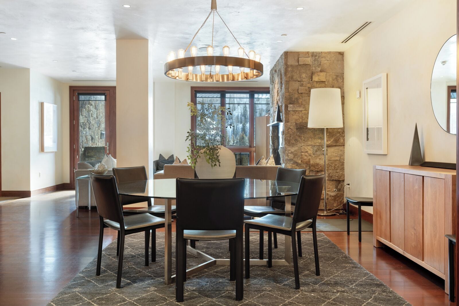A dining area with a round table, six chairs, a chandelier, a stone wall, and a large window.