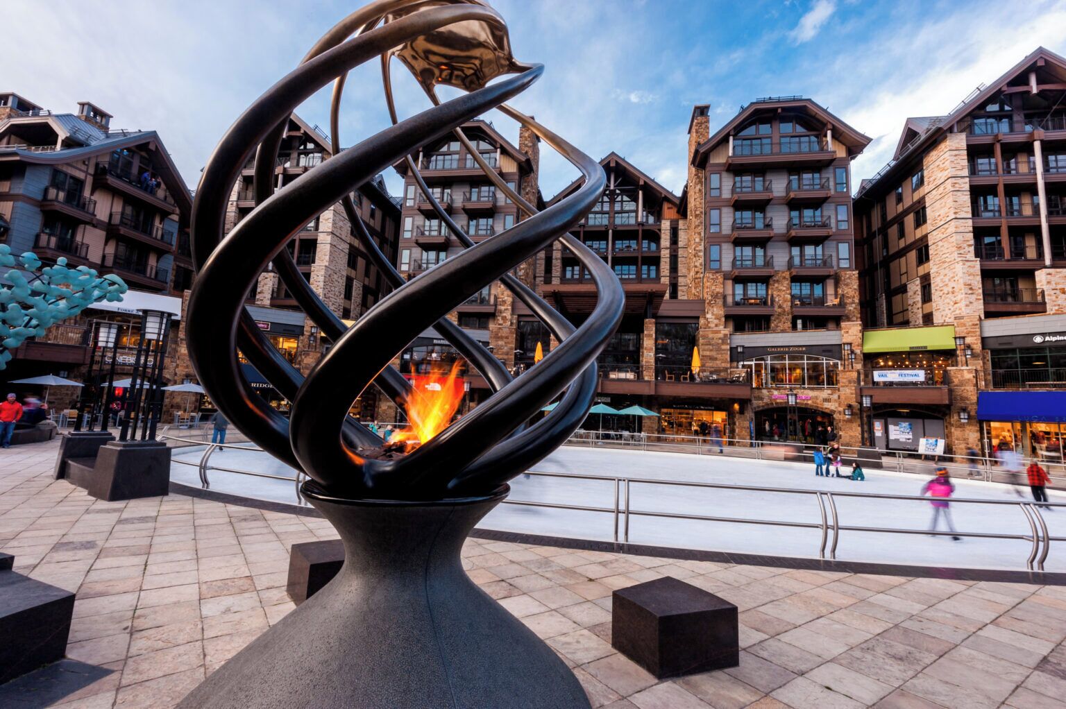 A modern sculpture with a fire in the center, set in a plaza with stone tiles, surrounded by multi-story buildings with balconies and storefronts.