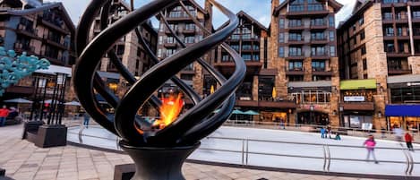 A modern sculpture with a fire in the center, set in a plaza with stone tiles, surrounded by multi-story buildings with balconies and storefronts.