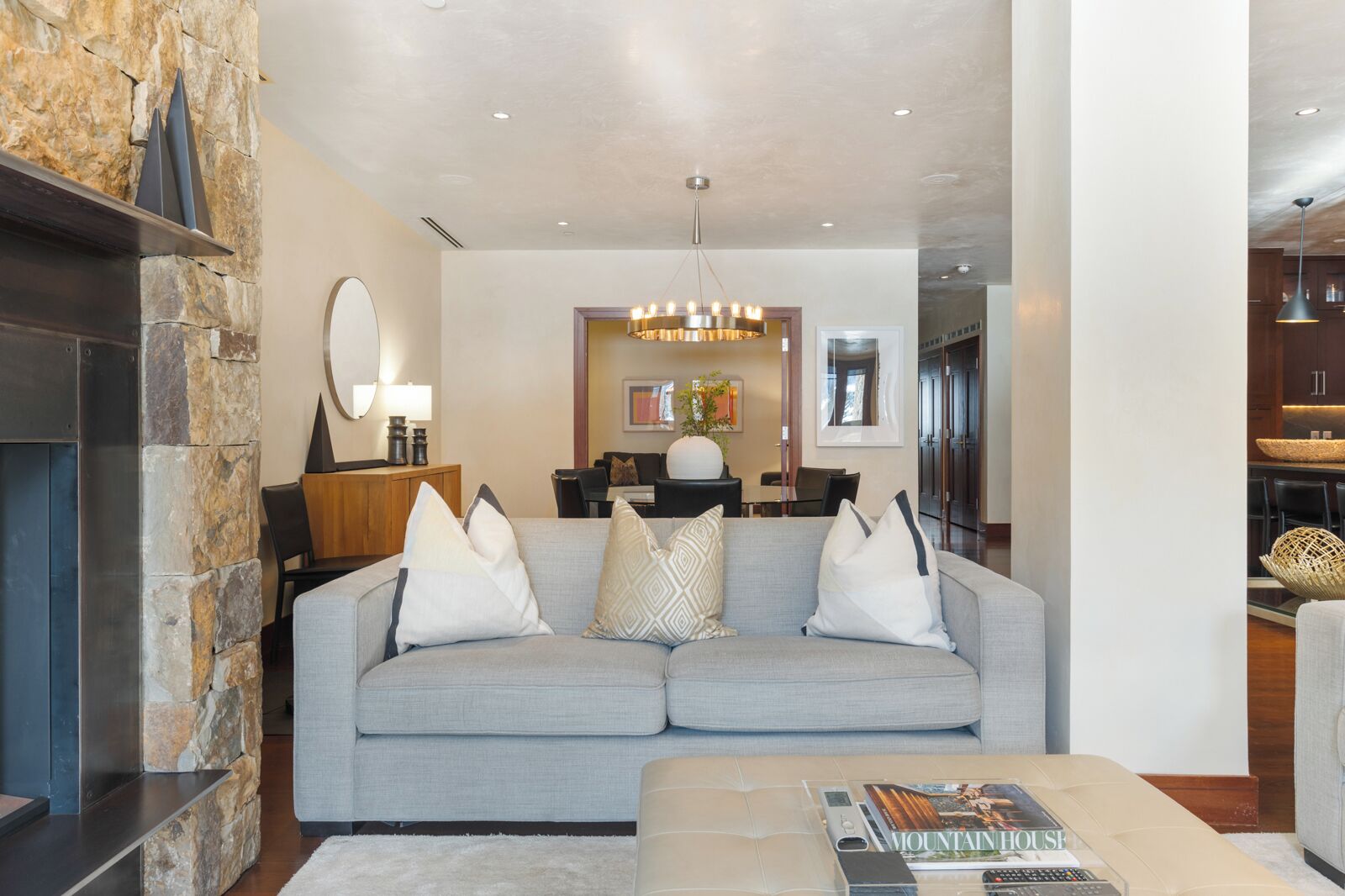 A modern living room with a stone accent wall, a grey sofa with patterned cushions, a coffee table with a magazine, and a dining area in the background.