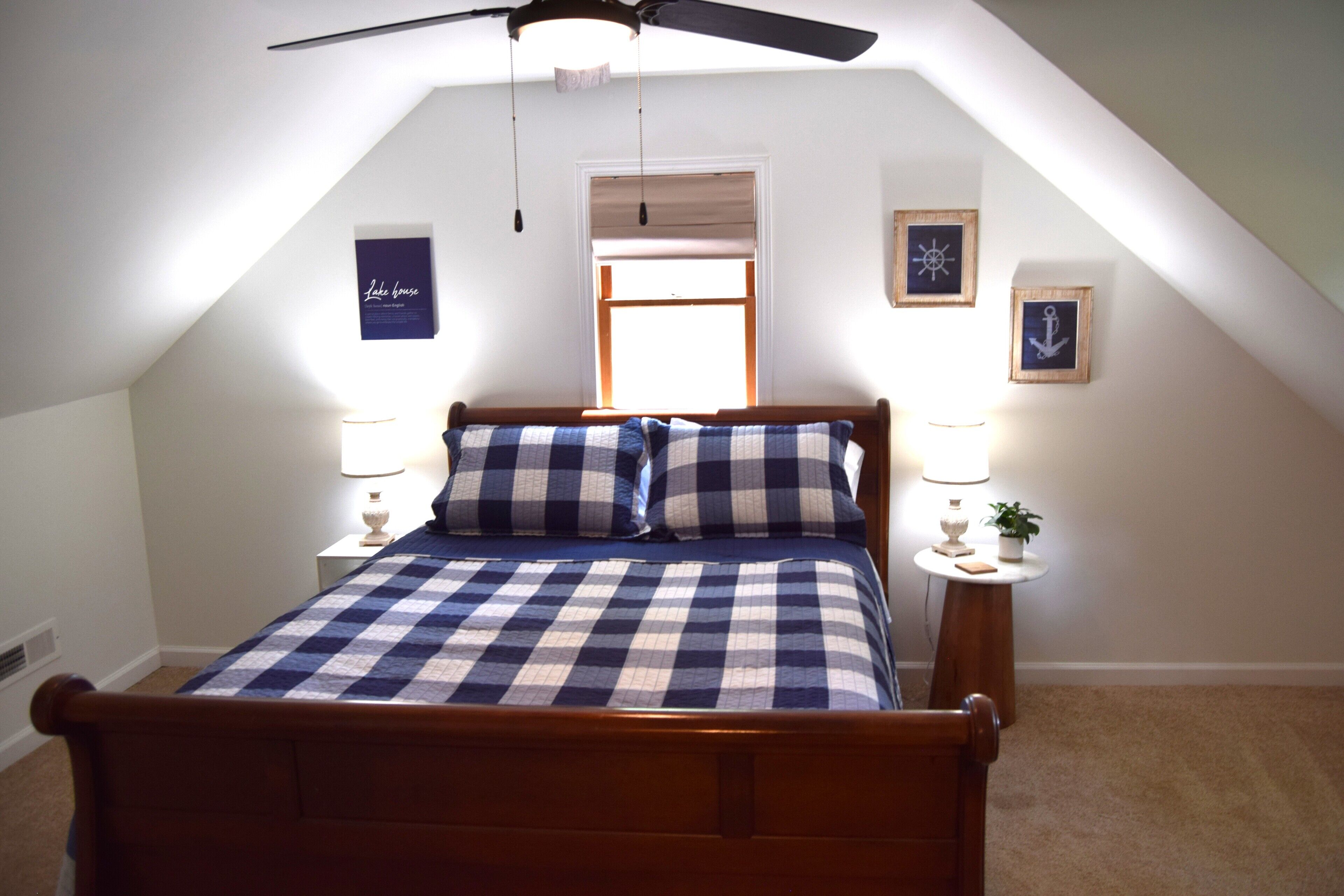 A bedroom with a sloped ceiling, a bed with checkered bedding, two bedside tables with lamps, and framed pictures on the walls.