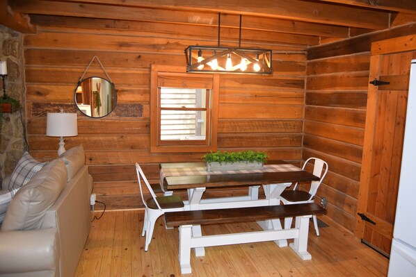 Interior de una cabaña de madera con un sofá blanco, una mesa de comedor de madera con bancos, un espejo colgante y una ventana con persianas.