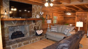 A cozy log cabin interior with a stone fireplace, a flat-screen TV mounted above it, a leather sofa, and a ceiling fan. - 3-bedroom cabin at Norris Lake, Sleeps 8 WiFi Lake access Covered boatslip (Speedwell)