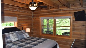 A wooden cabin bedroom with a bed, a ceiling fan, a window with a view of greenery, and a nightstand with a lamp. - 3-bedroom cabin at Norris Lake, Sleeps 8 WiFi Lake access Covered boatslip (Speedwell)