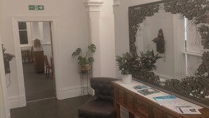 Une pièce avec un grand miroir, une coiffeuse en bois et un fauteuil en cuir foncé. Il y a une porte qui mène à une autre pièce et un panneau indiquant la sortie sur le mur.