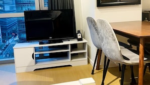 A modern living room with a flat-screen TV on a white entertainment unit, a grey upholstered chair, a wooden dining table, and a large window with a city view.