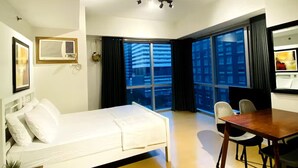 A hotel room with a bed, a dining table with chairs, a small table with a plant, a large window with curtains, and a ceiling light fixture.