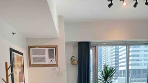 A modern bedroom with a large bed, a bedside table, a wall-mounted air conditioner, a window with curtains, and a view of buildings outside.