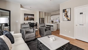 Un salon moderne avec un canapé, deux fauteuils, une table basse et un ventilateur de plafond. On y trouve également un coin repas avec une table et des chaises, et une cuisine à l’arrière-plan.