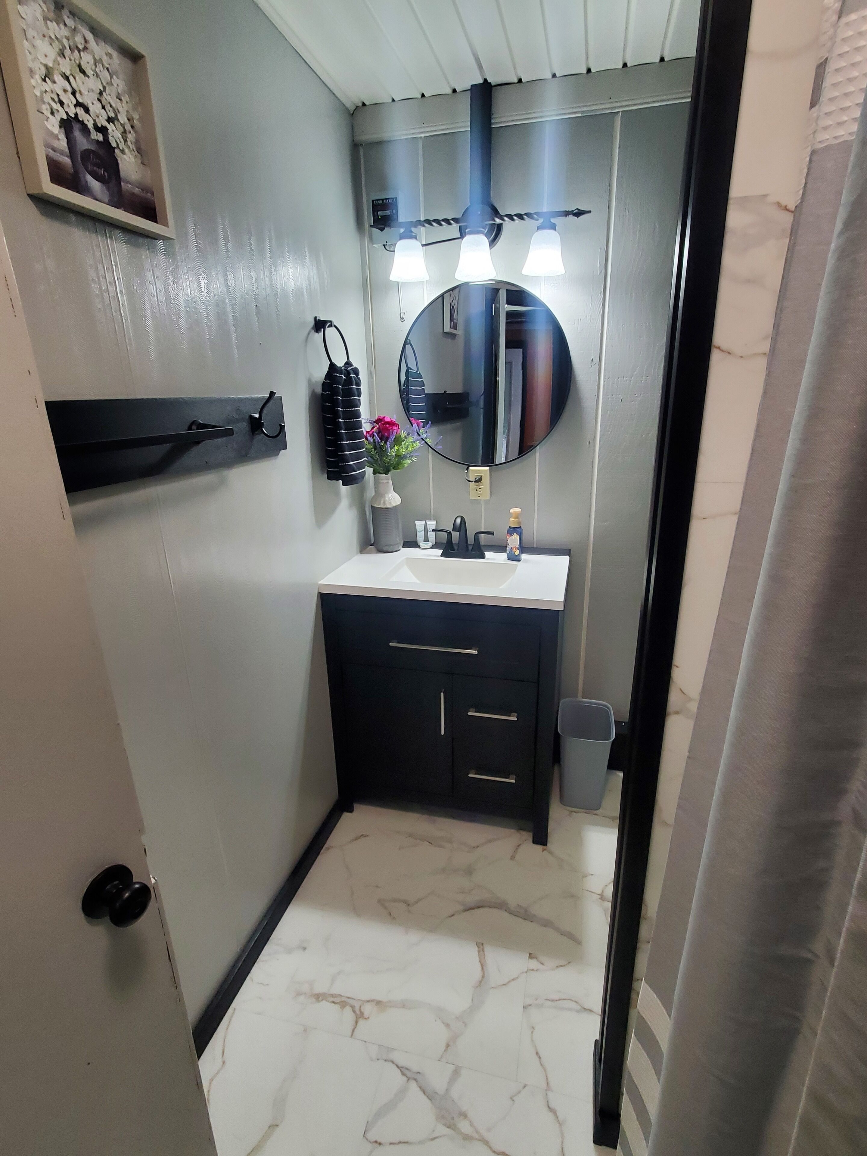 Une salle de bain avec un sol en marbre blanc, un lavabo noir surmonté d’un plan de travail blanc, un miroir rond au-dessus du lavabo et un porte-serviettes avec une serviette qui y est accrochée.