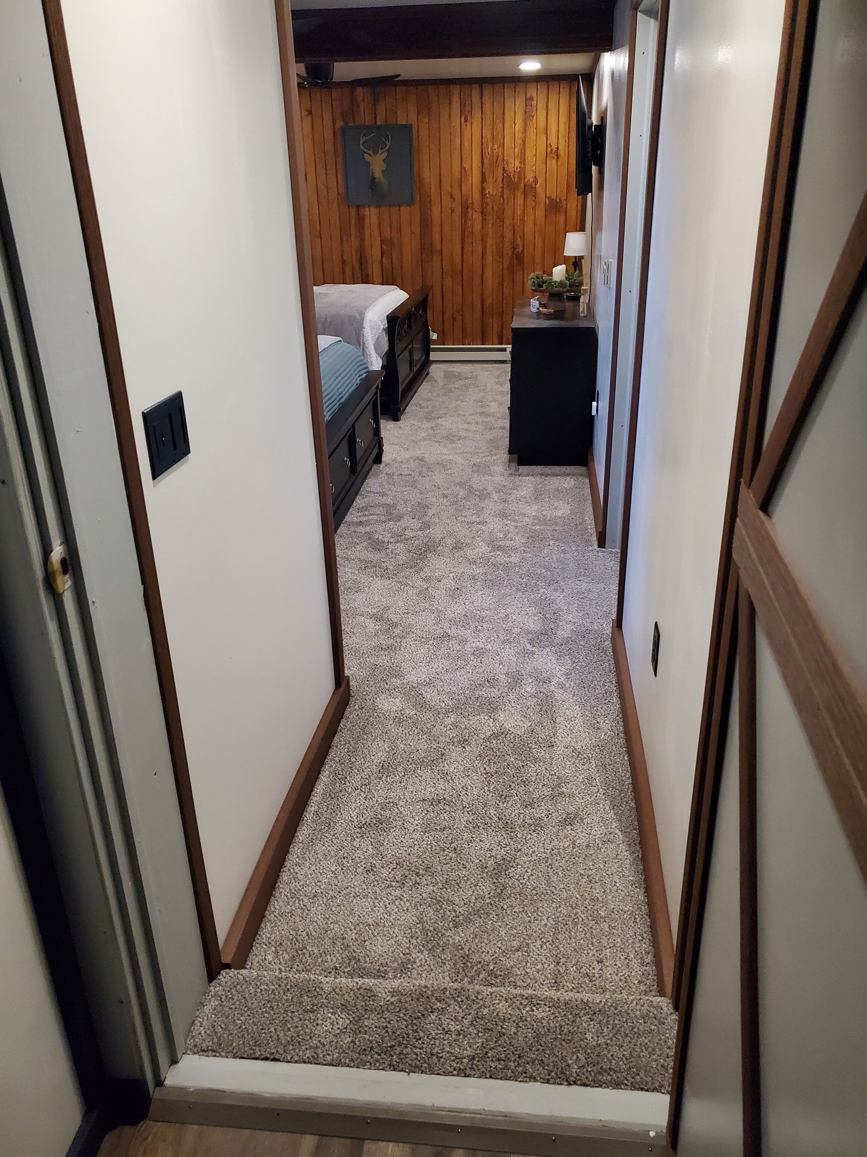 A narrow hallway with a carpeted floor leading to a bedroom with a bed and a dresser.