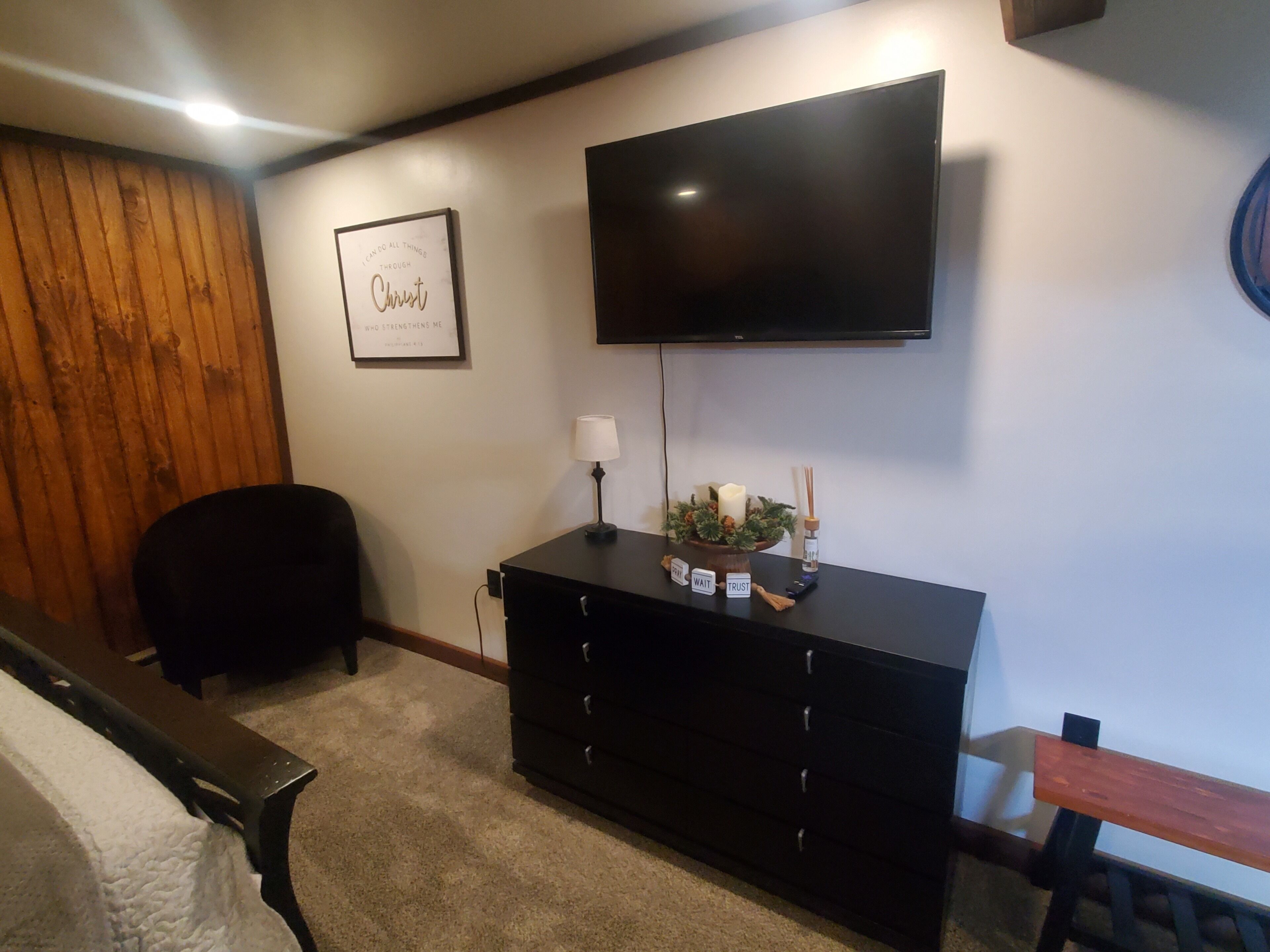 Quarto com centro de entretenimento preto, televisão de tela plana, quadro com citação inspiradora, pequena mesa com velas e uma cadeira.