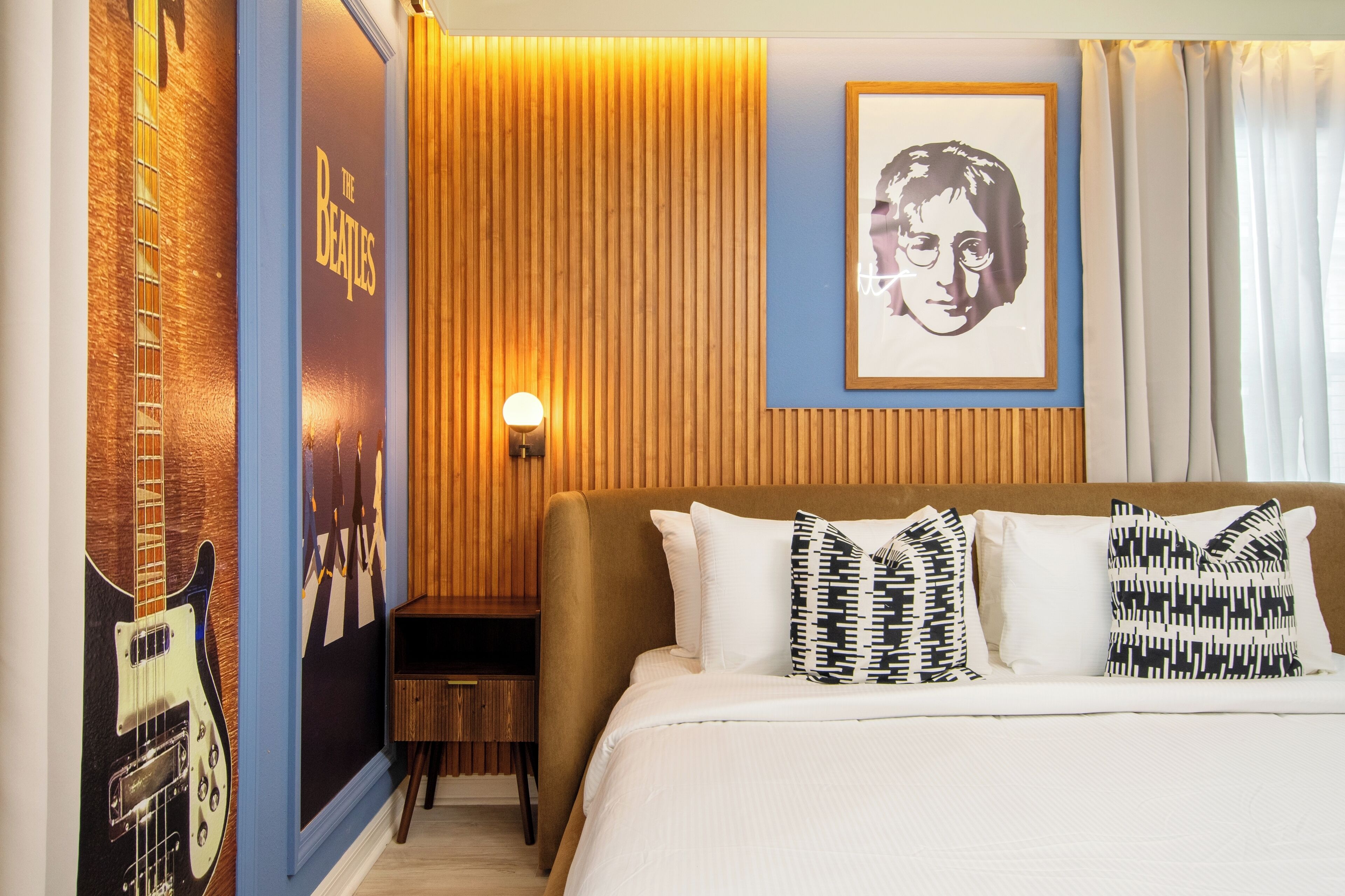 Habitación de hotel con una cama, una lámpara de pared, un cuadro enmarcado y un cartel de The Beatles.