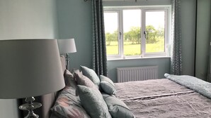 A bedroom with a bed, pillows, a lamp, a window with curtains, and a view of greenery outside.