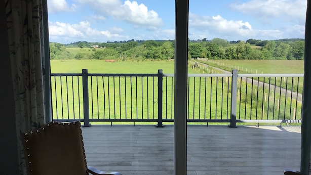 A view from a room with a wooden deck and a metal railing, overlooking a green field and a road with a fence.