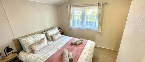 Un dormitorio con cama, almohadas, una mesita de noche, lámpara y ventana con cortinas.
