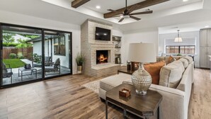 A spacious living room with a fireplace, a ceiling fan, and a large window overlooking a garden.