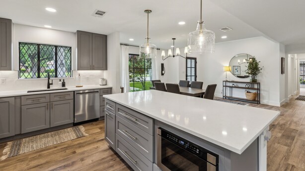 A modern kitchen with grey cabinets, a large island, stainless steel appliances, and pendant lighting.
