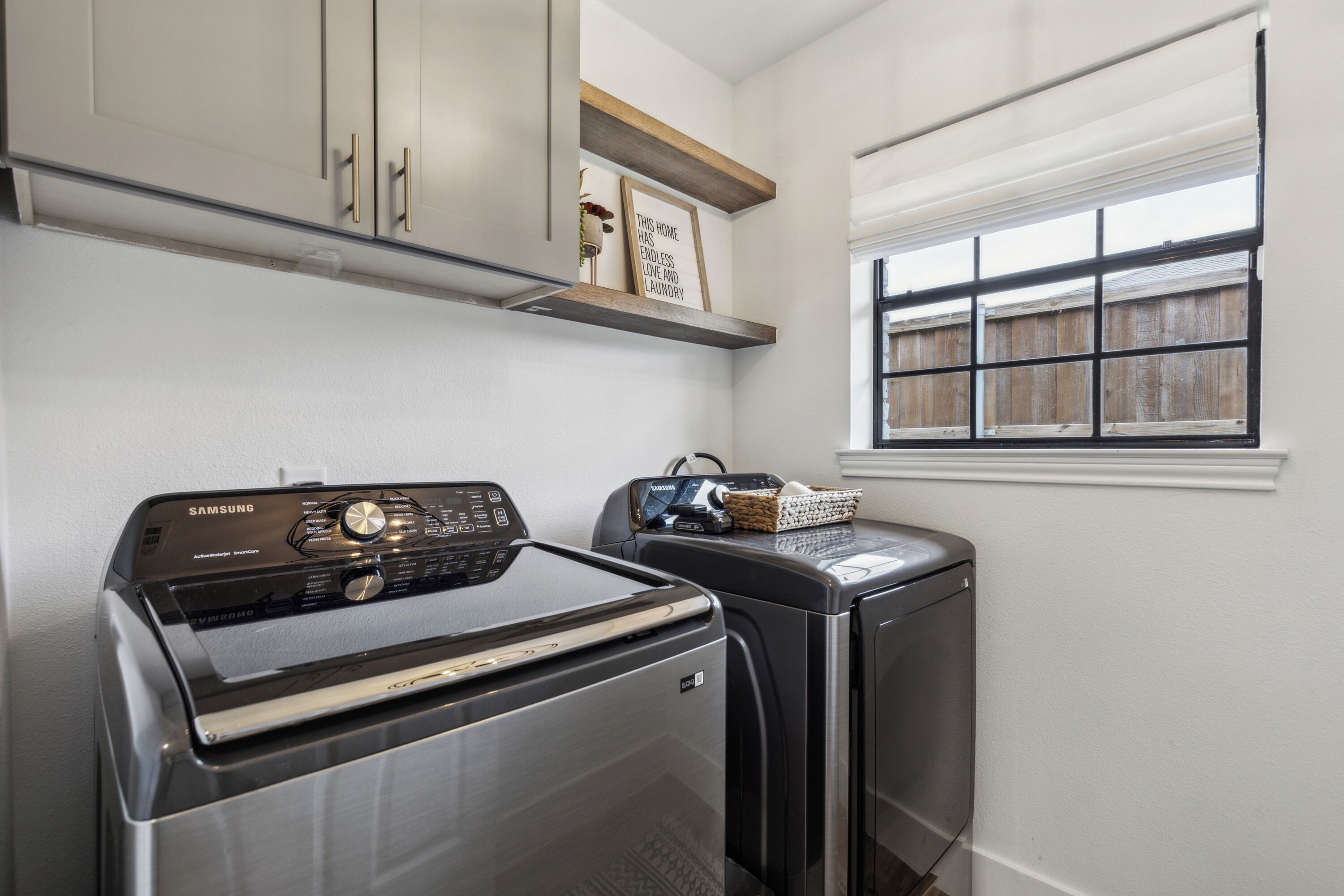 A laundry room with a Samsung washer and dryer, wooden cabinets, a window with blinds, and a shelf with a framed sign.