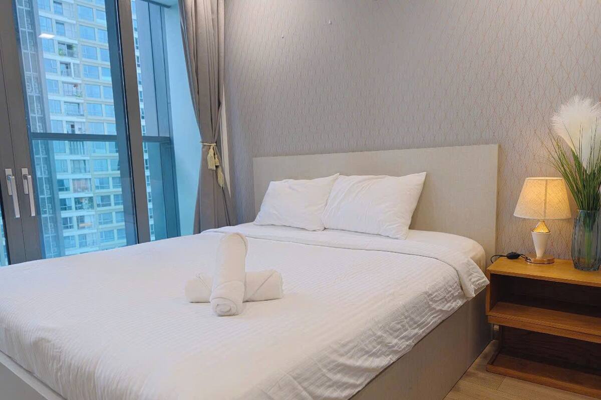 A neatly made bed with white linens and towels, a wooden nightstand with a lamp, and a view of a cityscape through large windows.