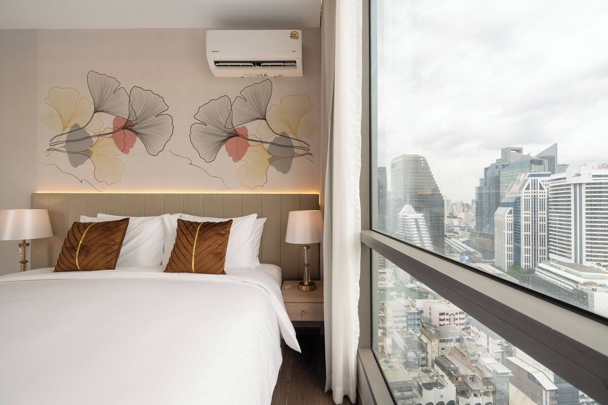 Une chambre d’hôtel avec un grand lit, deux tables de chevet, un climatiseur fixé au mur et une vue sur la ville par une grande fenêtre.