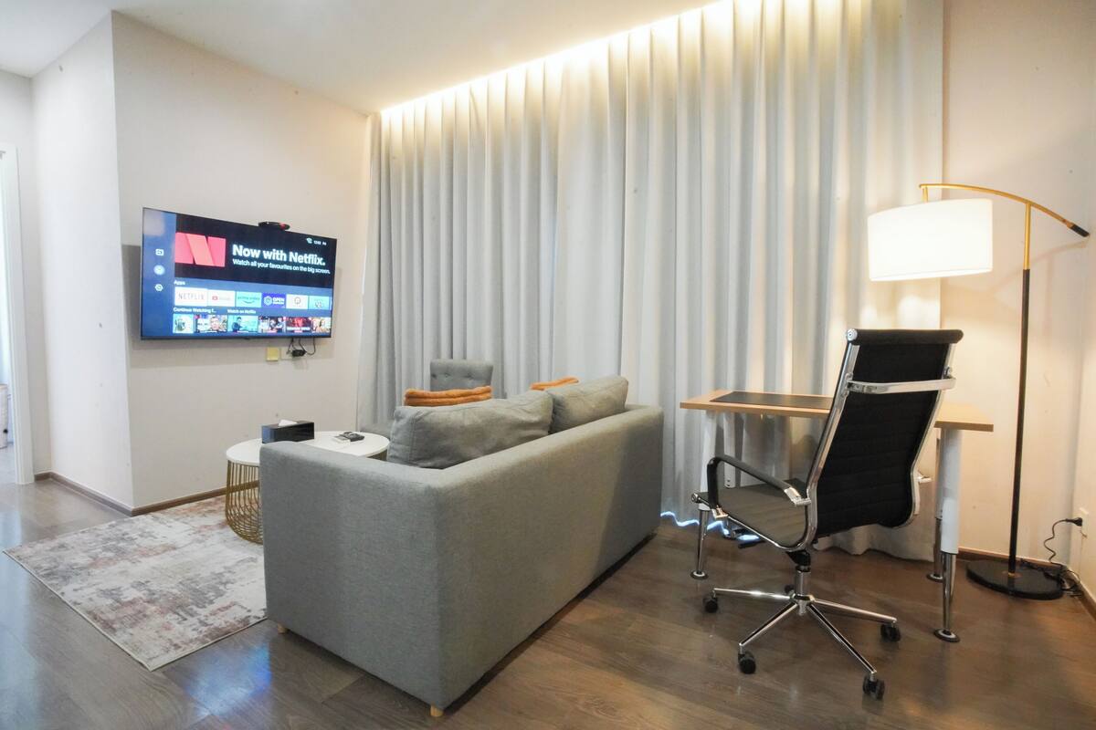 Une chambre d’hôtel moderne avec un canapé gris, un téléviseur fixé au mur, un bureau avec une lampe et une chaise.