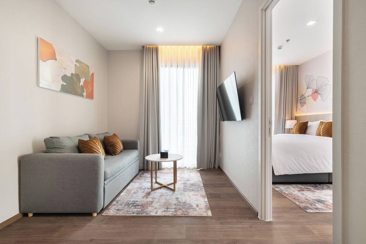 Une chambre d’hôtel moderne avec un canapé gris, une petite table ronde, un lit recouvert d’une couette blanche et un téléviseur fixé au mur.