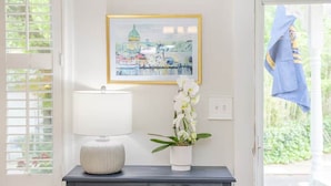 Une commode en bois foncé, sur laquelle sont posés une lampe blanche et un pot d’orchidée. Au-dessus de la commode, on peut voir un tableau encadré représentant un paysage urbain.